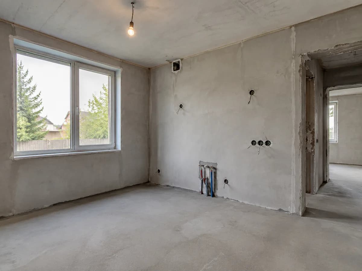 An empty room under renovation with bare plastered walls, a window, a doorway and pipes coming out of one wall