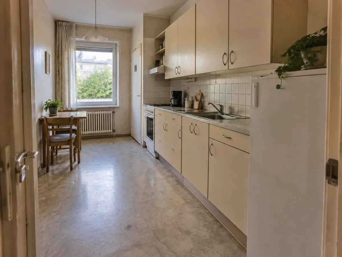 A narrow dated kitchen with beige cabinets along one wall, a small fridge and a dining table by the window